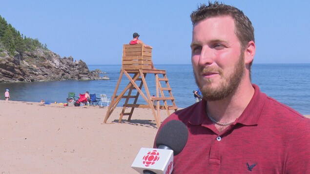 Un homme se fait interviewer sur une plage. Derrière lui, on voit la mer et un sauveteur. Un homme se fait interviewer sur une plage. Derrière lui, on voit la mer et un sauveteur.