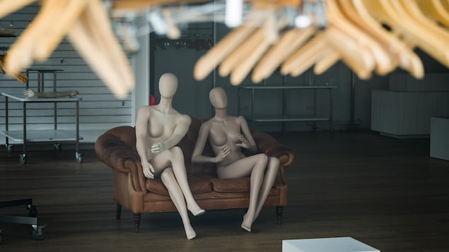 Deux mannequins assis sur un sofa brun au milieu d'un local vide, avec des cintres en avant-plan