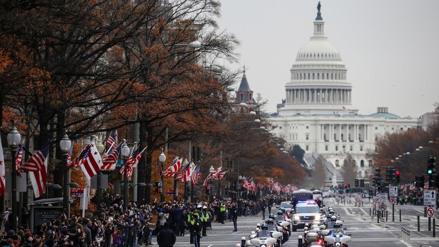 Un cortège funèbre défile dans une avenue aux trottoirs bondés de monde avec des drapeaux américains hissés. Un cortège funèbre défile dans une avenue aux trottoirs bondés de monde avec des drapeaux américains hissés.