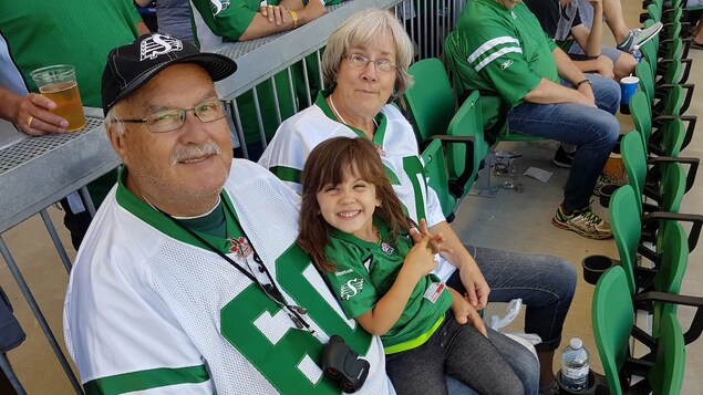 Butch Gullacher pose pour la caméra. À côté de lui se trouve sa femme Kathleen. Sur ses genoux est assise sa petite-fille Athéna. Ils sont à une partie de football et portent tous des chandails aux couleurs des Roughriders de la Saskatchewan.