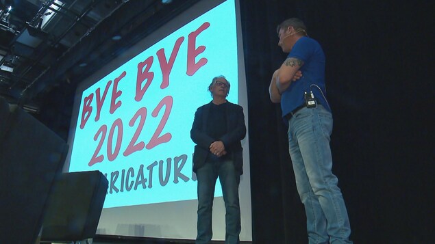 Deux hommes sur une estrade. Derrière eux, un écran géant sur lequel on peut lire Bye bye 2022 en caricatures.
