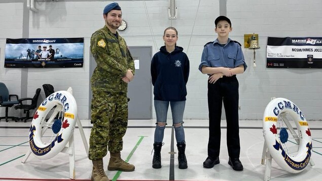 Un adulte, et une jeun adulte et un adolescent se tenant debout entre deux bouées de sauvetage représentant le Corps de Cadets de la Marine Royale Canadienne 117 Jacques-Cartier (CCMRC 117) Jacques-Cartier.
