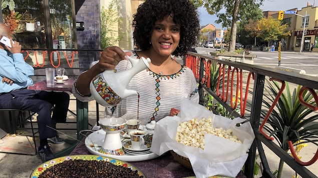 Une femme assise à la terrasse devant un homme qui parle au téléphone. Jebena à la main, elle verse le café dans des petites tasses rangées sur un plateau à coté de grains de café et de maïs soufflé.