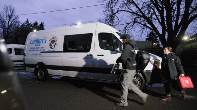 Une camionnette blanche au logo "CAHOOTS", le soir. À droite, deux personnes transportent des équipements.