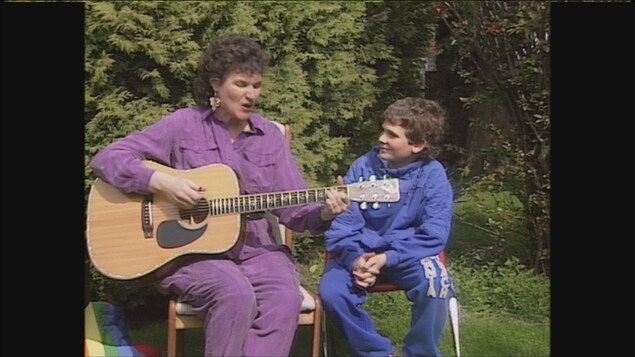 une dame assise dans un jardin qui chante et joue de la guitare et un garçon assis à côté d'elle.