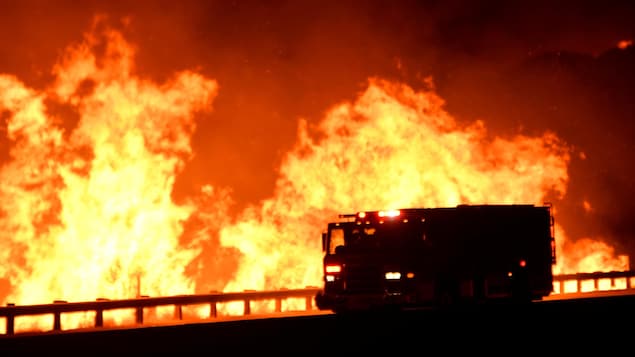 Un camion de pompier se déplace sur une route qui est à proximité d'un énorme incendie. Les flammes sont plus hautes que le véhicule.