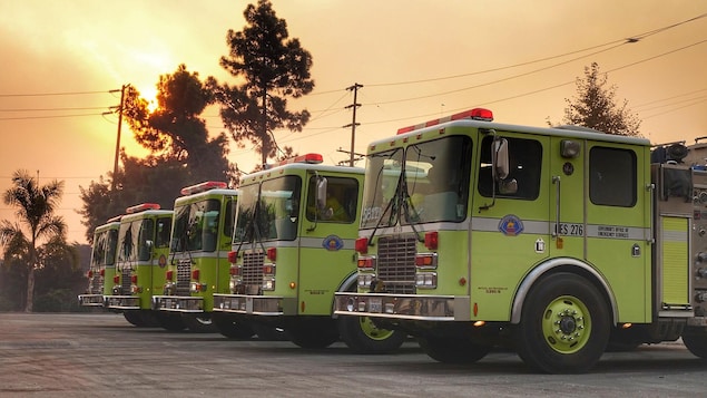 Cinq camions de pompiers alignés sous un ciel enfumé