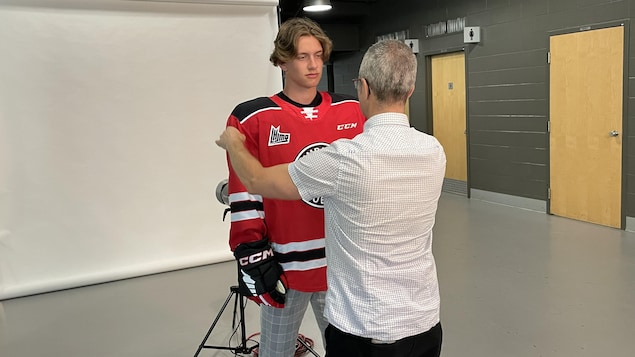 Un jeune homme porte un chandail de joueur de hockey rouge et noir.. Il est dans un studio de photo, prêt à se faire photographier. Le photographe lui ajuste le chandail.