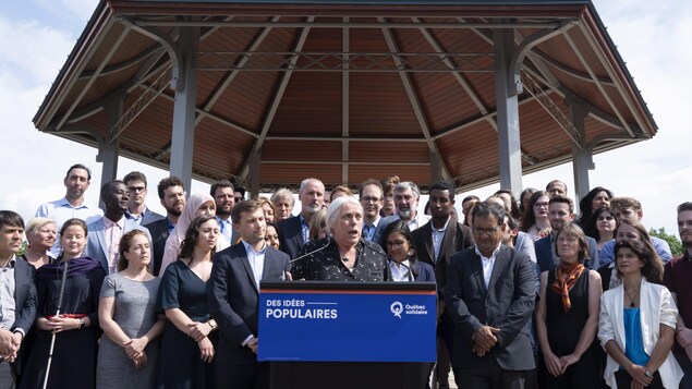 Manon Massé parle à un lutrin, derrière elle se trouvent quelques dizaines de candidats.