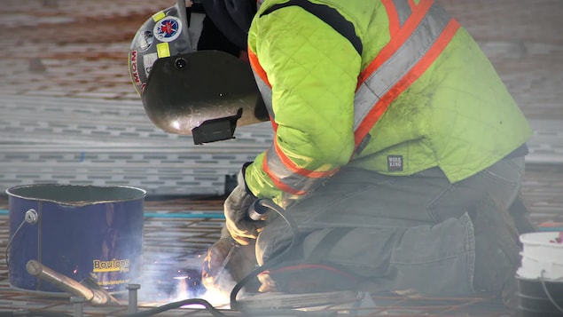 A construction worker is kneeling, wearing a protective helmet and holding a soldering device.