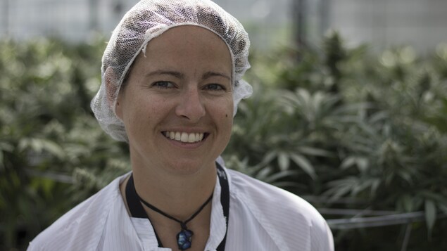 Une jeune femme portant un filet sur les cheveux et un sarrau blanc sourit à la caméra devant des plants de cannabis. Une jeune femme portant un filet sur les cheveux et un sarrau blanc sourit à la caméra devant des plants de cannabis.