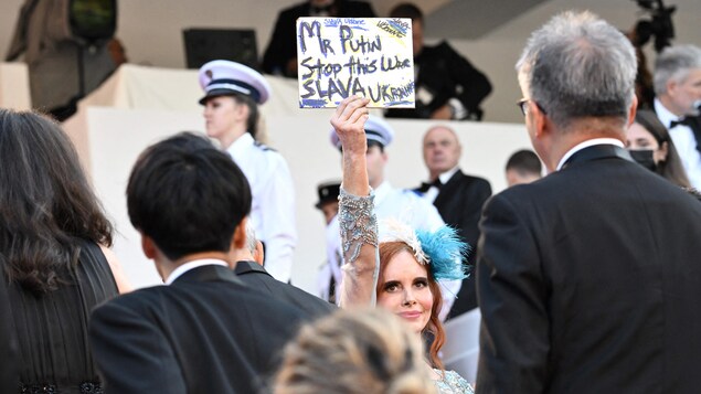 Une femme brandit un carton sur lequel est inscrit un message antiguerre sur les marches du palais des festivals à cannes, entourée d'une foule de gens qui marchent face à elle.  