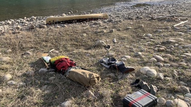 Des sacs étanches, un boîtier et  un canot son éparpillés sur la rive d'une rivière.