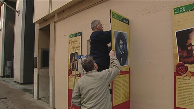 Deux hommes, dont un sur une échelle, retire un panneau de la Commission de la capitale nationale sur lequel on voit un portrait en noir et blanc de Lord Durham Deux hommes, dont un sur une échelle, retire un panneau de la Commission de la capitale nationale sur lequel on voit un portrait en noir et blanc de Lord Durham