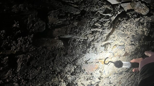 Un mur de pierre dans un couloir souterrain est illuminé partiellement par la lampe de poche que tient une personne.