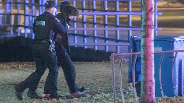 Deux policiers tiennent Carl Girouard par les mains, lors de son arrestation. Le suspect a les cheveux brun foncé et est vêtu de noir. 