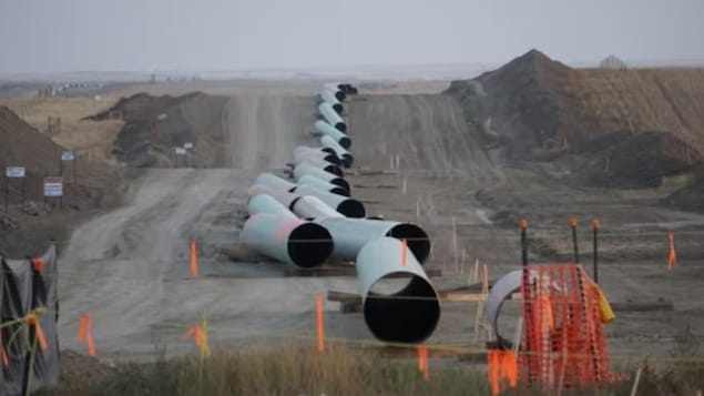 Une longue rangée de sections de pipelines de grand diamètre sont déposées bout à bout sur un terrain de construction sablonneux. 
