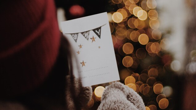 Une femme tient une carte de Noel devant un sapin.