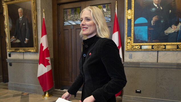 Catherine McKenna en mouvement, sourire aux lèvres. Elle se dirige face aux journalistes.