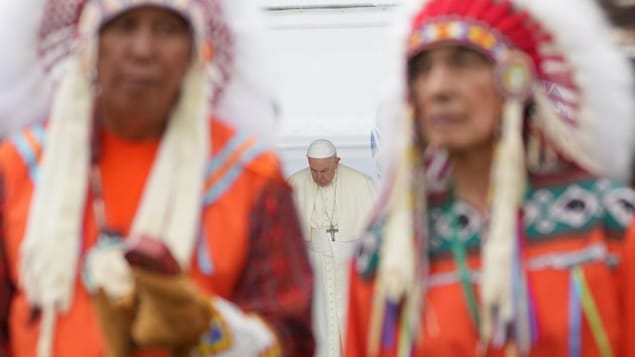 El Papa Francisco inclina la cabeza detrás de dos jefes indígenas.