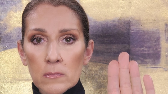 Céline Dion regarde la caméra, le visage sérieux. Elle lève sa main pour montrer le « D » qui y est inscrit.