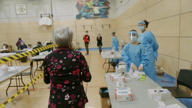 Dans un gymnase, une femme marche en direction de travailleuses de la santé munies d'équipements de protection individuelle.