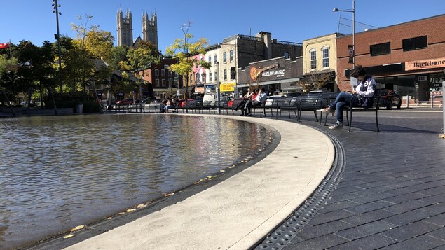 Des gens assis sur des bancs de parc devant une étendue d'eau avec les commerces de la rue principale derrière eux.