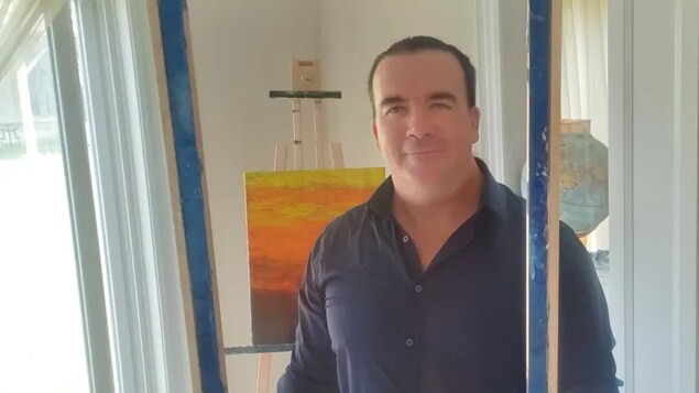 Chad Solomon avec une chemise bleue et une peinture en arrière plan. 