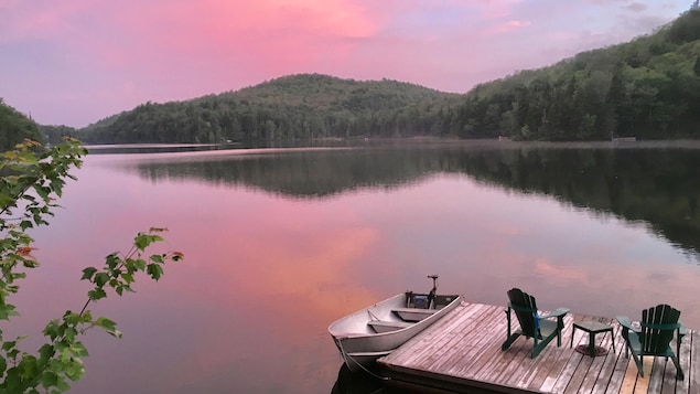 Un lac à la tombée du jour, avec un quai en bois, des chaises et une embarcation; au premier plan, le feuillage d'un arbre.