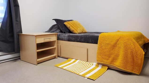 Une petite chambre à coucher avec un lit simple et des couvertures grise et jaune installées dessus.