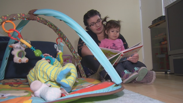 Une maman lit une histoire à une fillette pendant qu'un bébé joue sur un tapis d'éveil. Une maman lit une histoire à une fillette pendant qu'un bébé joue sur un tapis d'éveil.