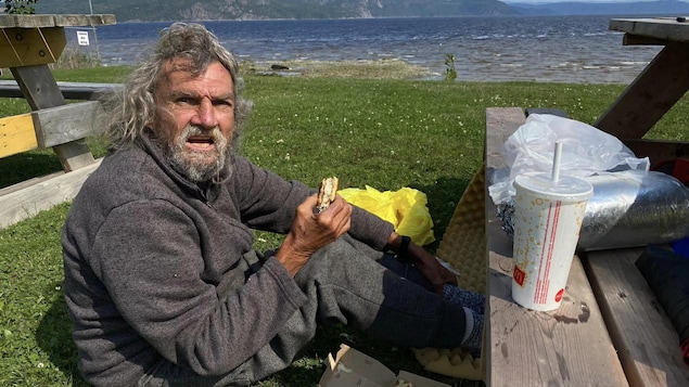 L'itinérant mange un hamburger assis sur le gazon sur les rives de la Baie des Ha! Ha!