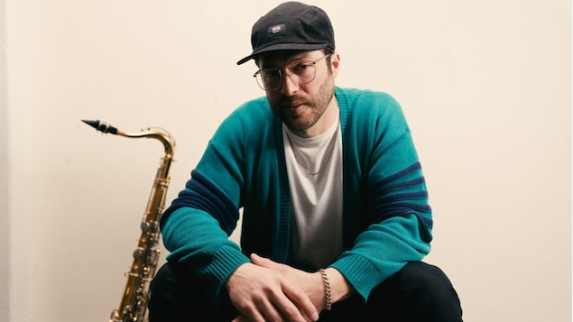 L’artiste Serge Paul assis, les mains croisées et un saxophone adossé sur un mur.