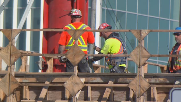 Des ouvriers sur un chantier de construction en plein travail. Ils portent un casque de protection sur la tête et un chasuble jaune ou orange.