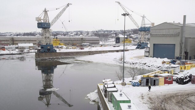 Une vue d'ensemble du chantier naval québécois Davie, en hiver, lors d'une journée grise. On y voit des grues et un travailleur qui marche près du fleuve.