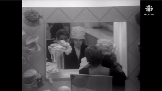Une femme essaie un chapeau devant un miroir pendant qu'une vendeuse assise derrière elle procède aux derniers ajustements.