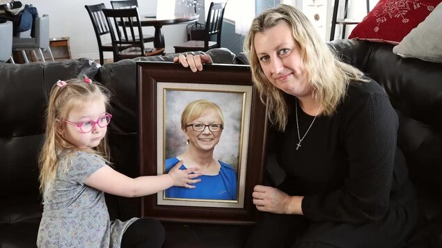 Une femme et une petite fille de 3 ans dans une résidence, assises sur un divan. Elles tiennent un cadre avec une photo d'une femme souriante, d'une soixantaine d'années, portant des lunettes et aux cheveux blonds coupés courts.