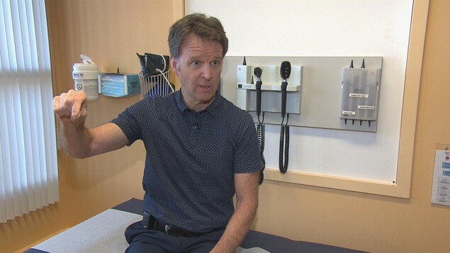 Gilles Morin fait le geste d'écrire un chèque assis sur une table de consultation dans un bureau médical. Gilles Morin fait le geste d'écrire un chèque assis sur une table de consultation dans un bureau médical.