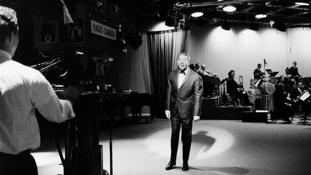Sur le plateau de l'émission, un caméraman film le chanteur Charles Trenet et derrière, l'orchestre.