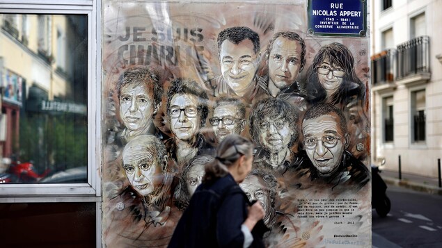 Une femme passe devant une murale reproduisant les visages de 10 employés de Charlie Hebdo.