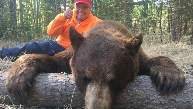 Un homme portant un pull orange fluo sourit à côté d'un gros ours mort et gisant sur un tronc d'arbre.