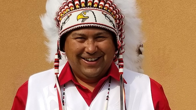Un homme portant une chemise rouge et une veste blanche porte une coiffe traditionnelle autochtone ainsi qu'un collier représentant un orignal. Un homme portant une chemise rouge et une veste blanche porte une coiffe traditionnelle autochtone ainsi qu'un collier représentant un orignal.