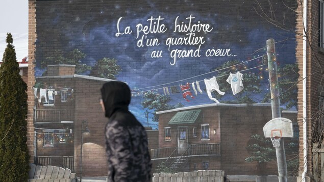  Une jeune qui passe devant une murale sur laquelle on peut lire : '' La petite histoire d'un quartier au grand coeur...''