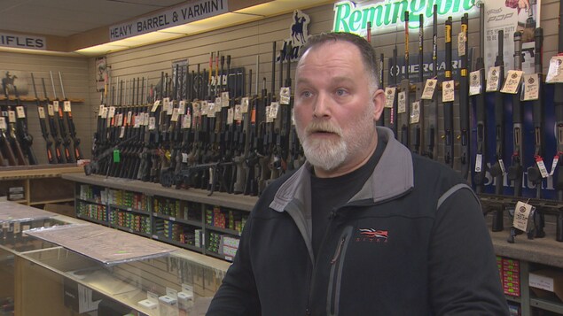 Un homme se tient devant un comptoir, derrière lui des armes sont exposées sur des étagères.