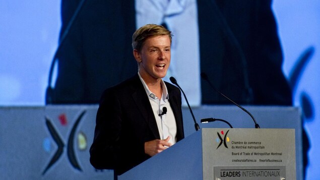 Une photo de Chris Hughes en train de donner un discours devant un écran géant rediffusant son image.