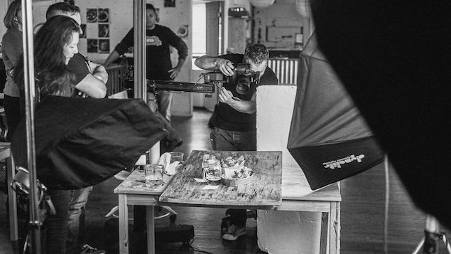 Photo en noir et blanc d'une session de photos en studio. Christian Lalonde est penché sur son objectif. Il fait une photo gastronomique avec des plats et deux bières dans des pots Masson avec poignées. L'équipe autour de lui le regarde travailler. Photo en noir et blanc d'une session de photos en studio. Christian Lalonde est penché sur son objectif. Il fait une photo gastronomique avec des plats et deux bières dans des pots Masson avec poignées. L'équipe autour de lui le regarde travailler.