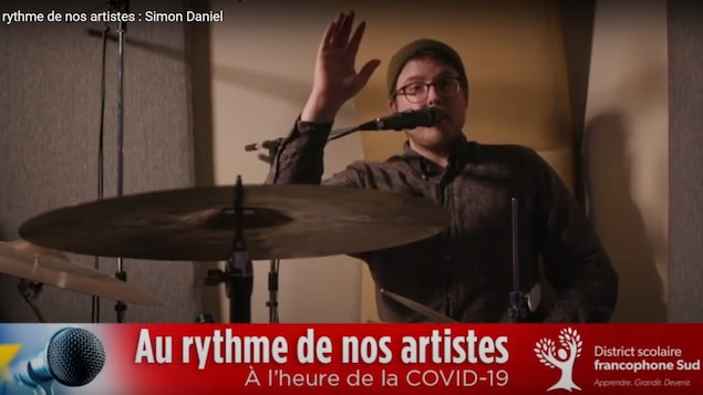 Simon Daniel assis à sa batterie, saluant la caméra.
