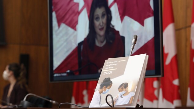 Chrystia Freeland apparaît sur un écran. En avant-plan se trouve le document de la mise à jour économique, sur un présentoir.