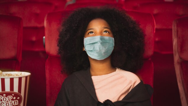Une jeune fille portant un masque de protection est assise dans une salle de cinéma, avec un popcorn.