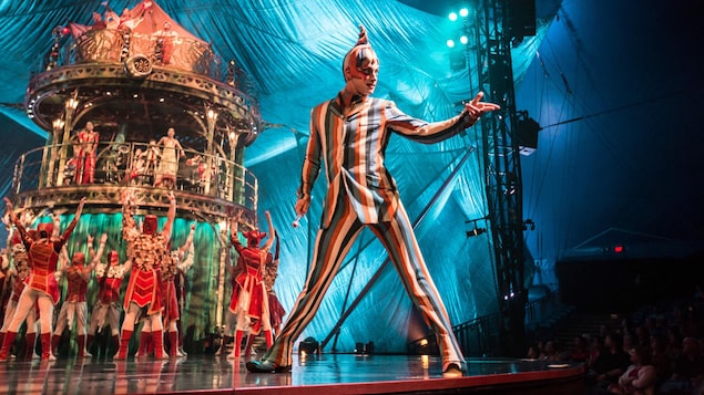Un artiste de cirque déguisé sur scène tend la main. Derrière lui, on voit un carrousel entouré par plusieurs personnes.